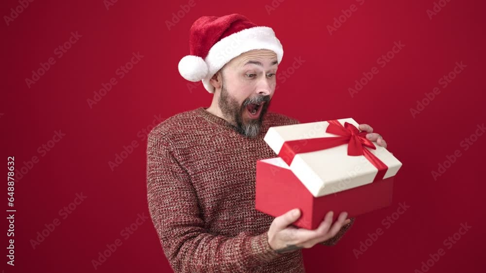 Young bald man wearing christmas hat unpacking gift over isolated red background Young bald man wearing christmas hat unpacking gift over isolated red background