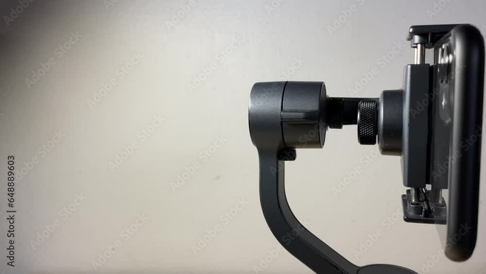 Black automatic gimbal stabilizer holding black phone on table against white wall indoor. with copy space. Technology and electronic gadget concept Black automatic gimbal stabilizer holding black phone on table against white wall indoor. with copy space. Technology and electronic gadget concept