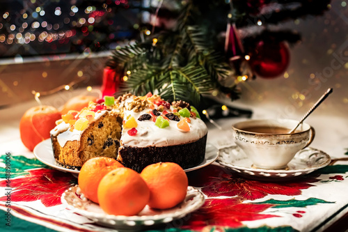 Christmas stollen with glaze, candied fruits and nuts on a saucer with a cup of tea, tangerines near a small Christmas tree.