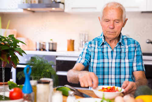 Elderly man cuts vegetables for salad at the table in the kitchen