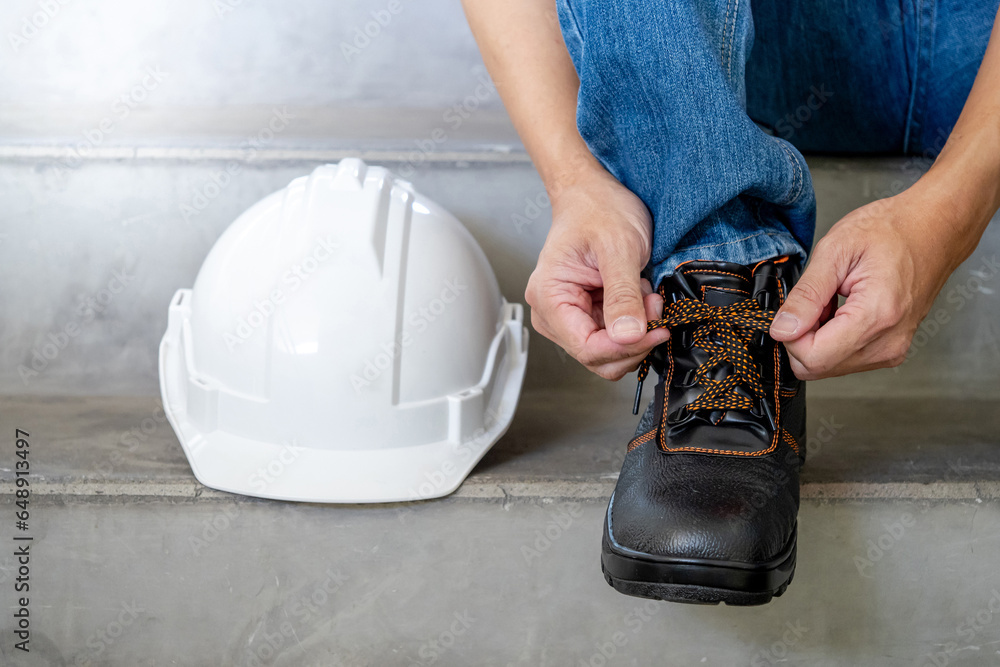 Male worker hands tying shoelaces on leather safety shoes with white protective helmet or hard hat on concrete stair in construction site. Safety workwear for worker and foreman - obrazy, fototapety, plakaty 