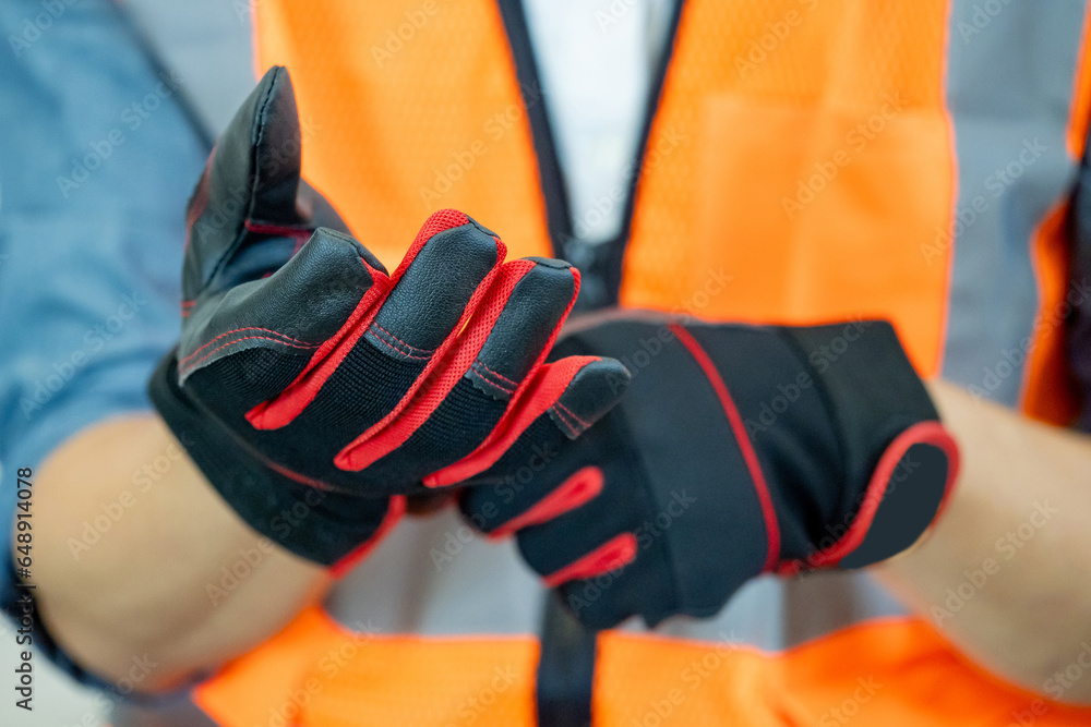 Male construction worker with reflective orange vest putting black and red protective gloves on his hands. Workwear for builder, foreman or engineer - obrazy, fototapety, plakaty 