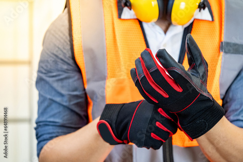 Male construction worker with reflective orange vest and yellow ear muffs putting black and red protective gloves on his hands. Workwear for builder, foreman or engineer photo