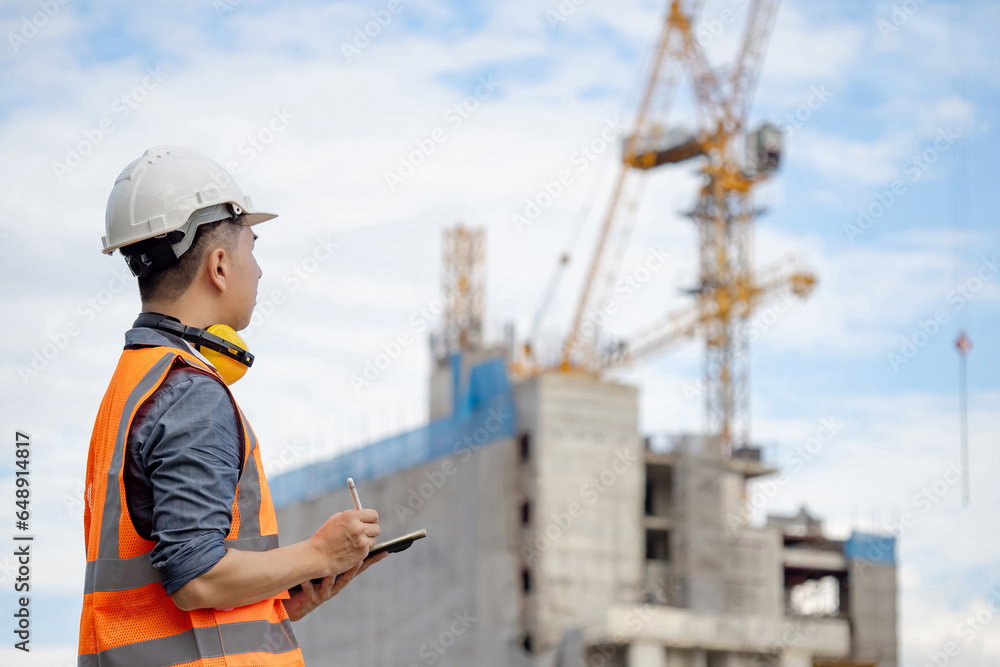 Male site engineer or foreman using digital tablet doing construction site inspection. Asian worker man with orange reflective vest and safety helmet looking at building structure and tower crane Male site engineer or foreman using digital tablet doing construction site inspection. Asian worker man with orange reflective vest and safety helmet looking at building structure and tower crane