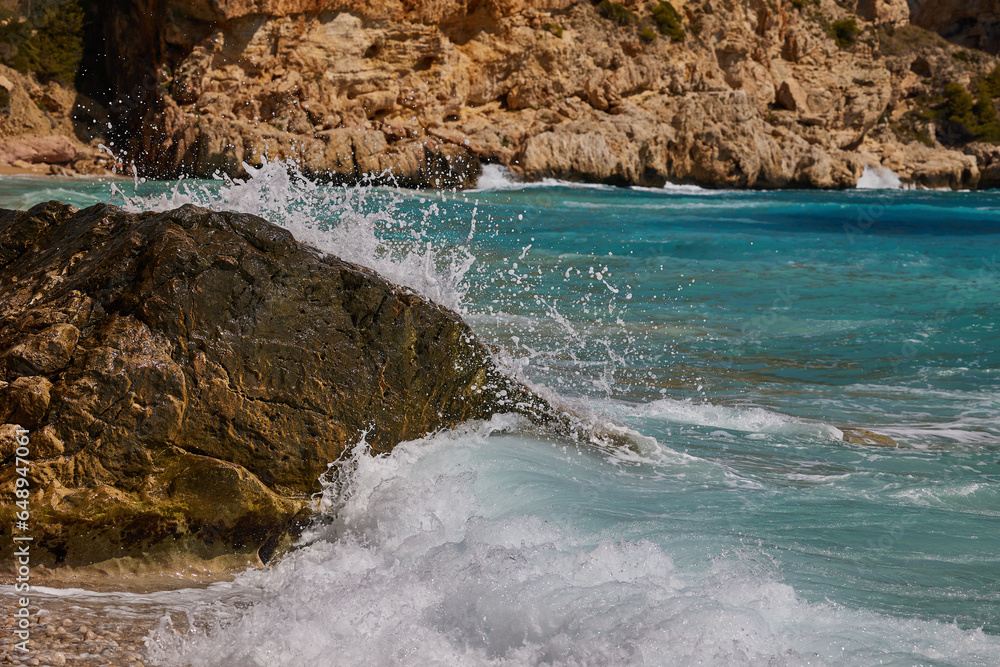 Waves waves crashing against rocks on the shores of the Mediterranean Sea during the day. Cala del Moraig beach in Benitachell of Alicante, Spain.