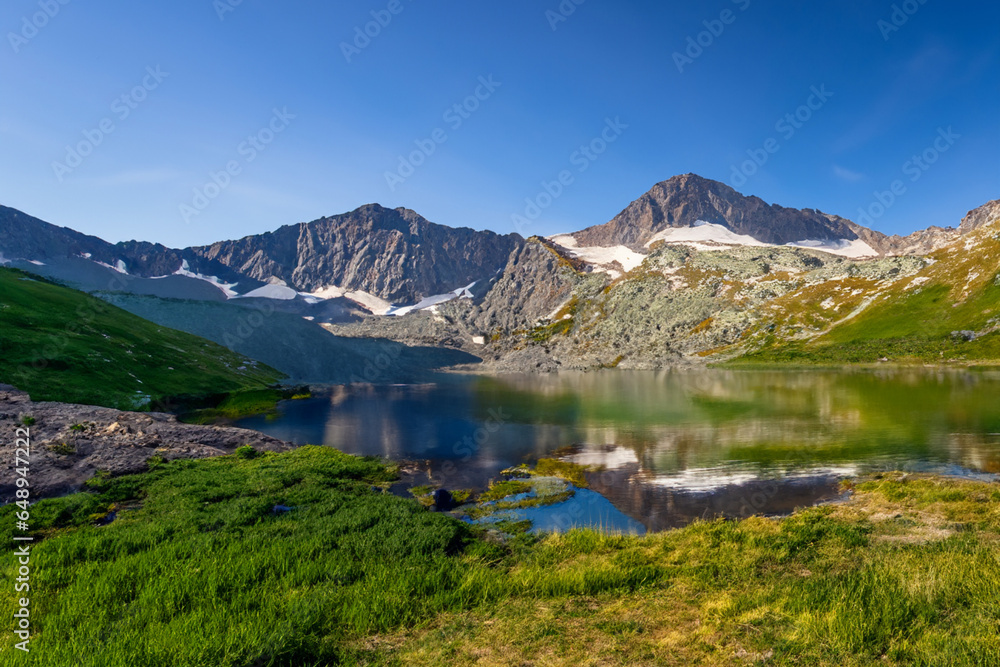 Obraz premium A serene mountain lake reflects the towering peaks and clear blue sky above, creating a tranquil and picturesque scene. The summer sun casts a warm glow, enhancing the beauty of the landscape.