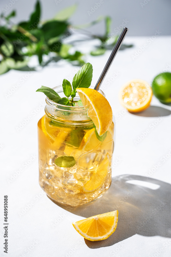 Fototapeta premium Lemonade drink or cold tea with fresh lemon, mint and ice in a jar. Refreshing citrus mojito cocktail on a light background with fresh fruits and shadows. Homemade healthy and detox drink concept