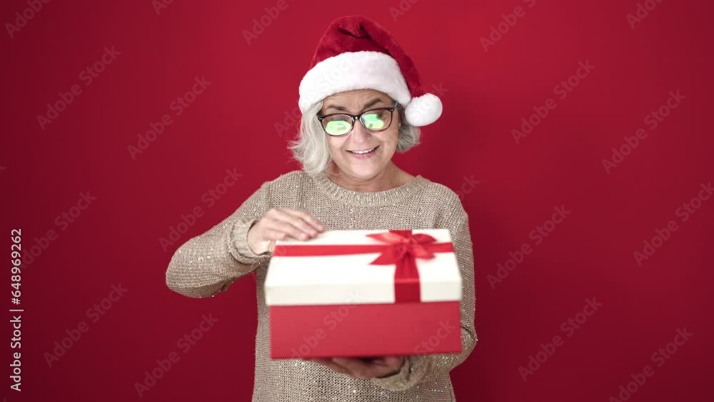 Middle age woman with grey hair wearing christmas hat unpacking gift over isolated red background Middle age woman with grey hair wearing christmas hat unpacking gift over isolated red background