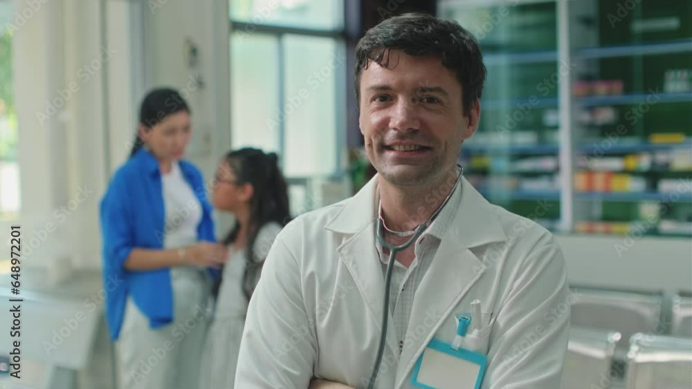 Portrait of cheerful pediatrician with stethoscope standing in clinic with woman and sick girl in background, looking at camera Portrait of cheerful pediatrician with stethoscope standing in clinic with woman and sick girl in background, looking at camera