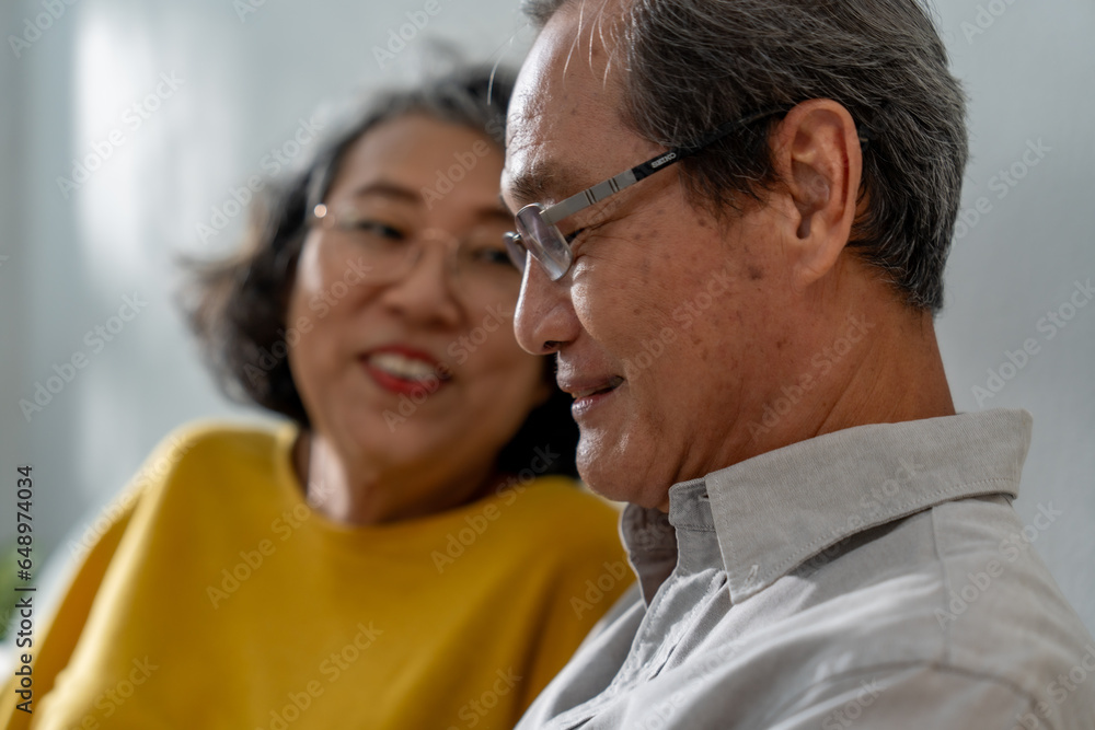 Senior Asian couple sitting happily relaxing on the sofa, using tablet devices with hands, smiling and laughing together.