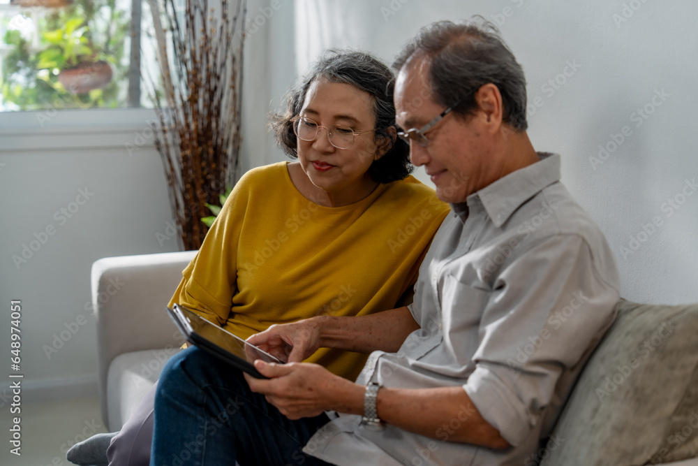 Senior Asian couple sitting happily relaxing on the sofa, using tablet devices with hands, smiling and laughing together. - obrazy, fototapety, plakaty 