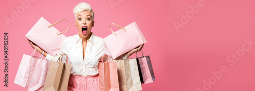 A middle-aged hipster woman holding shopping bags, surprised face with mouth open, pastel pink background copyspace, low prices sale and discounts for black friday concept