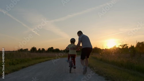 Dad teaches daughter to ride a bike. Happy family childhood dream concept of parent and little kids learn to ride a bike silhouette in the park.