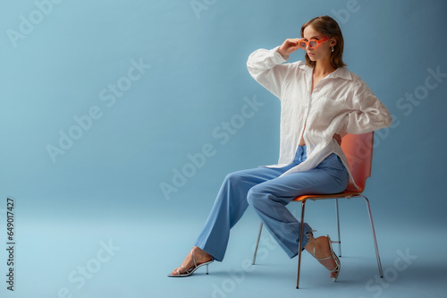  Fashionable young woman wearing trendy orange sunglasses, white linen shirt, trousers, metallic strap sandals, posing on blue background. Full-length studio portrait. Copy, empty space for text