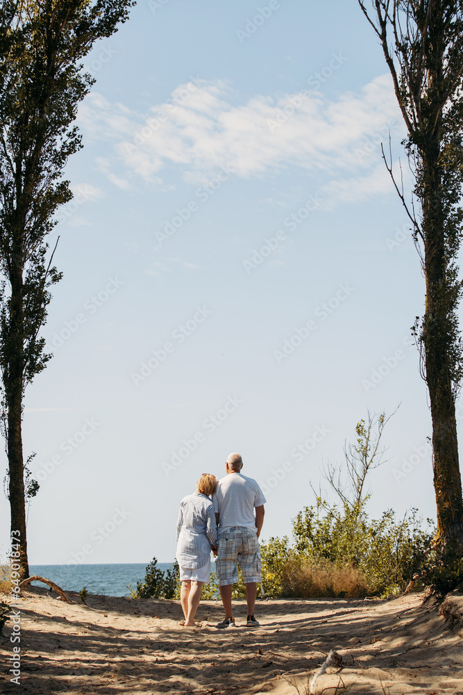 Happy elderly couple hold hands, stand among the trees, look at the sea. Happy elderly couple hold hands, stand among the trees, look at the sea.