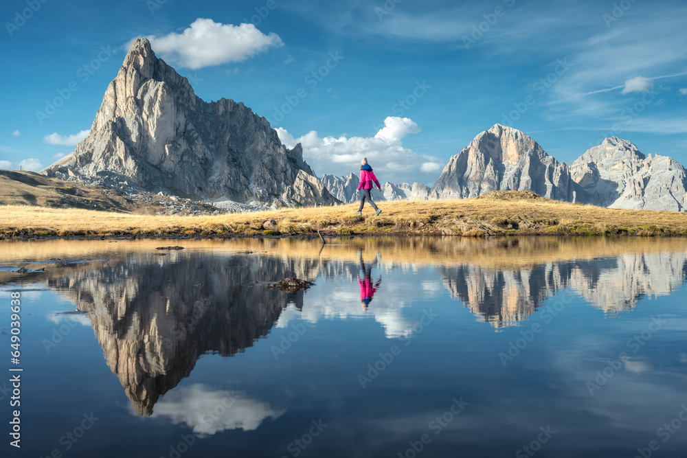 Walking woman and mountains reflected in lake at sunset in autumn in Dolomites, Italy. Standing girl on the shore of lake is looking on high rocks and blue sky with clouds in fall. Reflection in water - obrazy, fototapety, plakaty 
