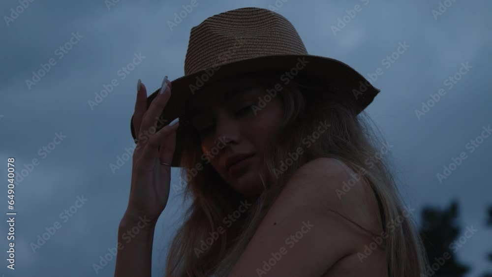 A young girl in a hat poses for the camera in a field against the backdrop of a beautiful summer sunset and looks into the camera. Slow motion. Close-up A young girl in a hat poses for the camera in a field against the backdrop of a beautiful summer sunset and looks into the camera. Slow motion. Close-up