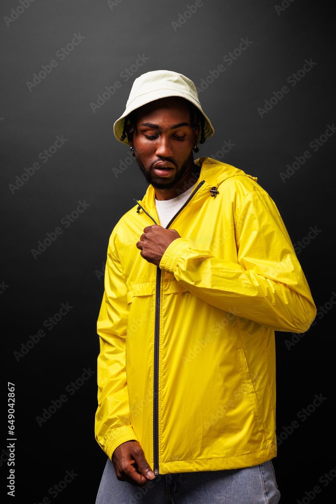 Trendy young african american man in yellow windbreaker jacket and bucket hat posing isolated on black Trendy young african american man in yellow windbreaker jacket and bucket hat posing isolated on black