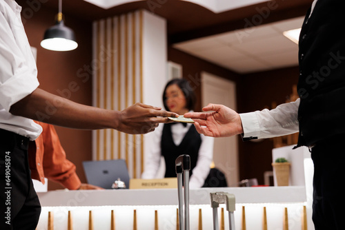 Guest giving cash to bellboy in lobby, paying money for luxury service to carry luggage and suitcases. Hotel concierge receiving payment from tourist, helping with trolley bags. Close up.