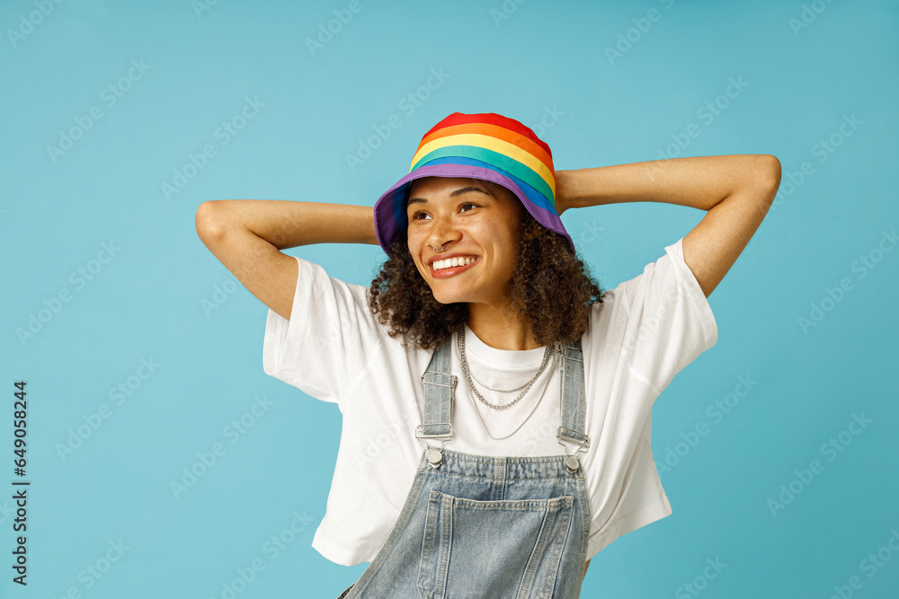 Naklejka premium Cheerful woman in rainbow cap standing with raised hands and looks away on blue studio background