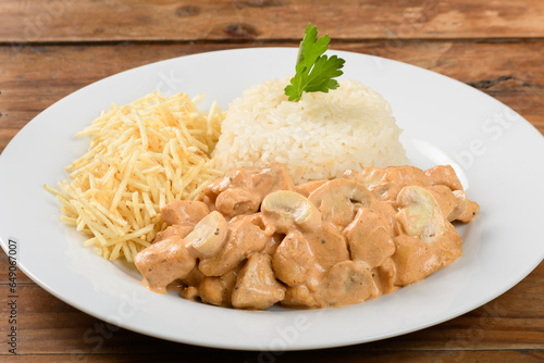 chicken stroganoff on a white plate with straw potatoes and rice, wooden table
