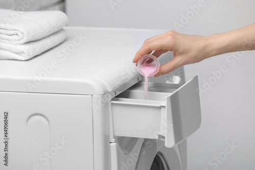Woman pouring fabric softener from cap into washing machine on light grey background, closeup