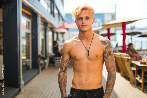 Beautiful attractive young man with blonde hair and body tattoos. Enjoying outdoors at a lively boardwalk lined with shops and restaurants, emphasizing a carefree spirit