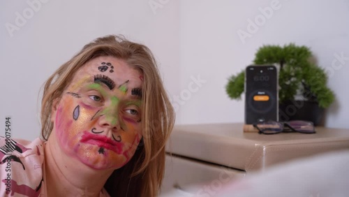 Smartphone alarm clock rings on bedside table, swollen disheveled woman wakes up, raises her head, smeared cosmetics, face painted with markers Hangover, morning after stormy party, bachelorette photo