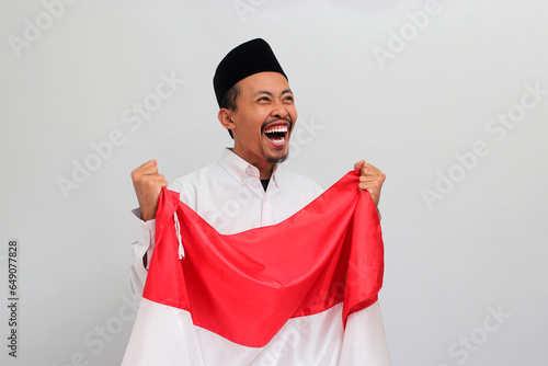 an Excited young Indonesian man, wearing a songkok, peci, or kopiah, is holding an Indonesian flag to celebrate Indonesia Independence Day on 17 August, isolated on white background photo