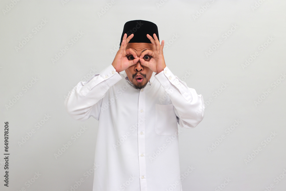 Excited young Indonesian man, wearing a songkok, peci, or kopiah, makes the OK sign gesture with his fingers, acting as binoculars with his eyes peeking through them, isolated on white background