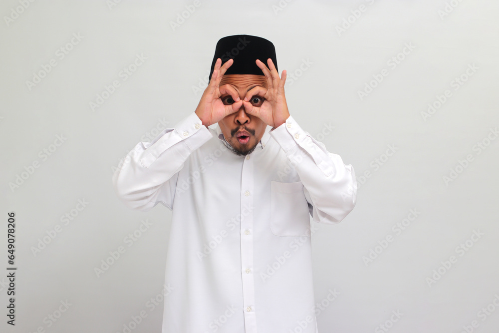 Excited young Indonesian man, wearing a songkok, peci, or kopiah, makes the OK sign gesture with his fingers, acting as binoculars with his eyes peeking through them, isolated on white background Excited young Indonesian man, wearing a songkok, peci, or kopiah, makes the OK sign gesture with his fingers, acting as binoculars with his eyes peeking through them, isolated on white background