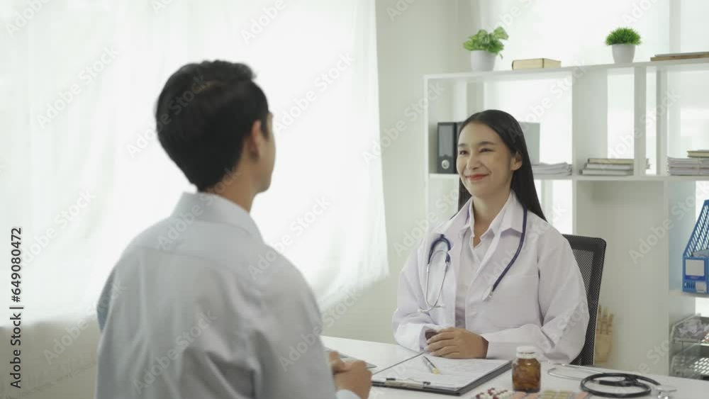 Young Asian doctor in white medical uniform uses clipboard to discuss results or symptoms with male patient sitting at table in clinic to discuss health issues. Young Asian doctor in white medical uniform uses clipboard to discuss results or symptoms with male patient sitting at table in clinic to discuss health issues.