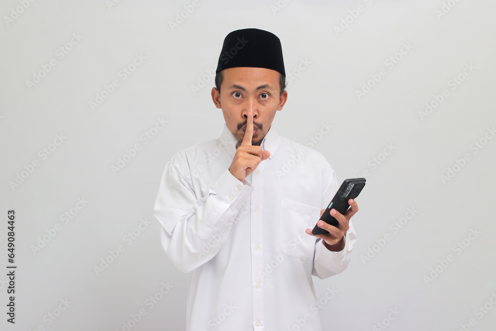 Young Indonesian man, wearing a songkok, peci, or kopiah, holding smartphone, fingers covering lips, making silent gesture, isolated on white background Young Indonesian man, wearing a songkok, peci, or kopiah, holding smartphone, fingers covering lips, making silent gesture, isolated on white background