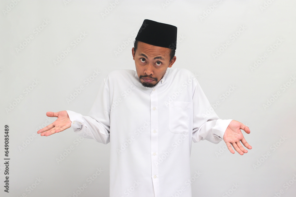 Confused young Indonesian man, wearing a songkok, peci, or kopiah, is shrugging his shoulders and looking at the camera with a puzzled expression, isolated on a white background Confused young Indonesian man, wearing a songkok, peci, or kopiah, is shrugging his shoulders and looking at the camera with a puzzled expression, isolated on a white background