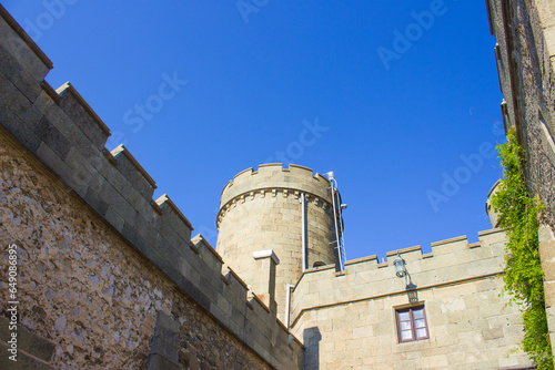 Photo of an old castle tower with a wall against the backdrop of mountains, deep sky and trees. Tower of an old castle against a background of blue sky and trees.