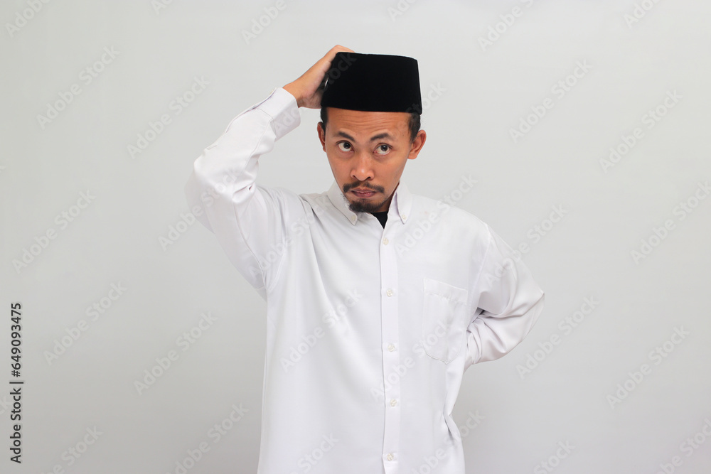 Pensive young Indonesian man, wearing songkok, peci, or kopiah, is scratching his head while looking at the camera, deep in thought, trying to find a solution, isolated on white background