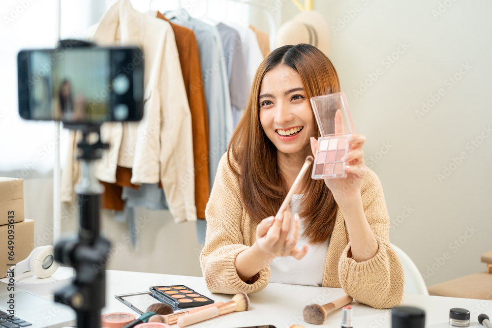 Young beautiful asian woman vlogger live broadcasting cosmetic makeup tutorial content with eye shadow looking at cell phone. Influencers professional beauty make up cosmetics record video on social - obrazy, fototapety, plakaty 