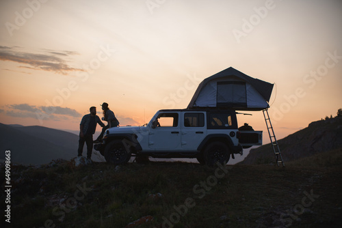Silhouette of couple of travelers in love with car against backdrop of sunset in mountains.
