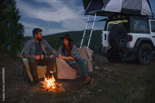 Сouple in love, man and woman on camp in mountains, sit by fire against backdrop of car in mountains.