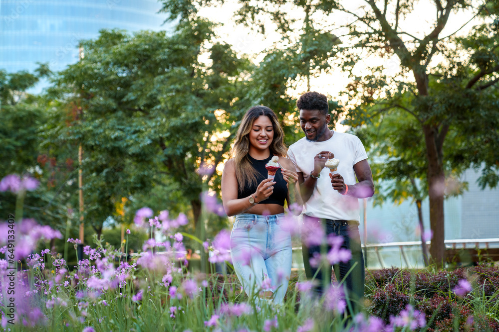 A beaming Afro-American man and a Latin woman, both in casual urban attire, savoring ice cream while strolling in a lush, flowery park at sunset. A beaming Afro-American man and a Latin woman, both in casual urban attire, savoring ice cream while strolling in a lush, flowery park at sunset.