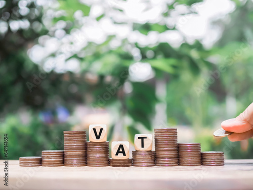 Close up hand putting coin in stack of coins, Wooden blocks with the word VAT. The concept of save money for paying vat in the future