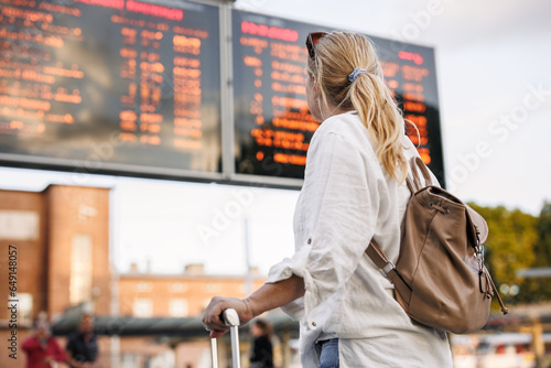 Woman passenger with backpack is checking arrival and departure board schedule timetable at train or bus station. Travel on vacation photo