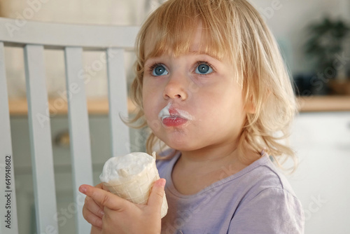 Baby girl enjoying ice cream. Pretty little toddler eating an ice-cream indoors, at home. Dining room background. Small child eats plombir and cream messy on her mouth. Cute kid with tasty sweet food. photo