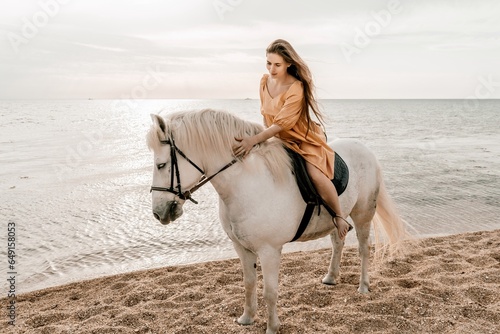 A woman in a dress stands next to a white horse on a beach, with the blue sky and sea in the background.