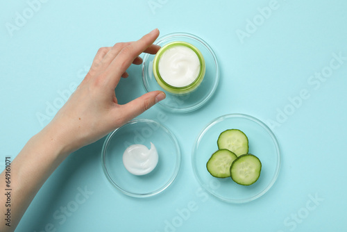 Jar with cream, sliced cucumber and cream on glass, female hand on blue background, top view