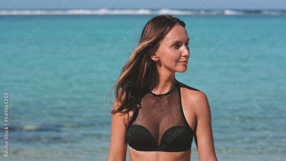 Sexy woman in bikini walking tropical beach coastline. Slim tanned brunette in black swimsuit against crystal blue ocean water on tropical sea shore during summer vacation lifestyle. Slow motion