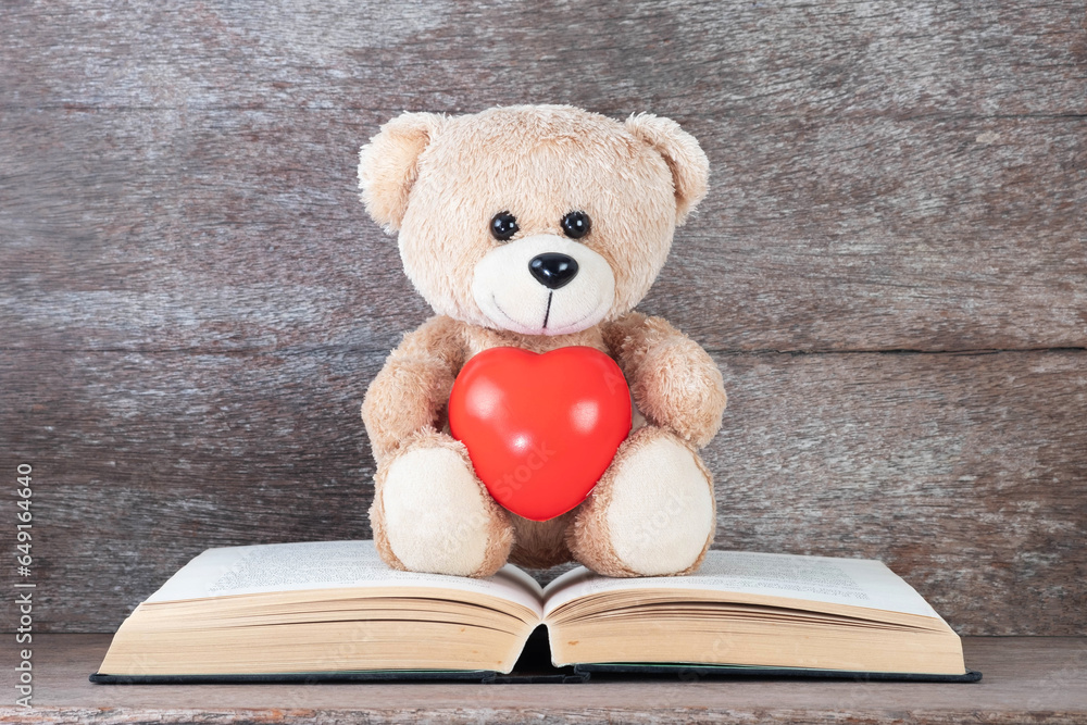 Teddy bear holding red heart sitting on old book and wooden background. Concepts of education, love and health. Teddy bear holding red heart sitting on old book and wooden background. Concepts of education, love and health.