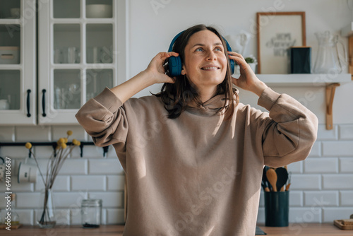 Happy woman wearing wireless headphones and listening to music in kitchen photo