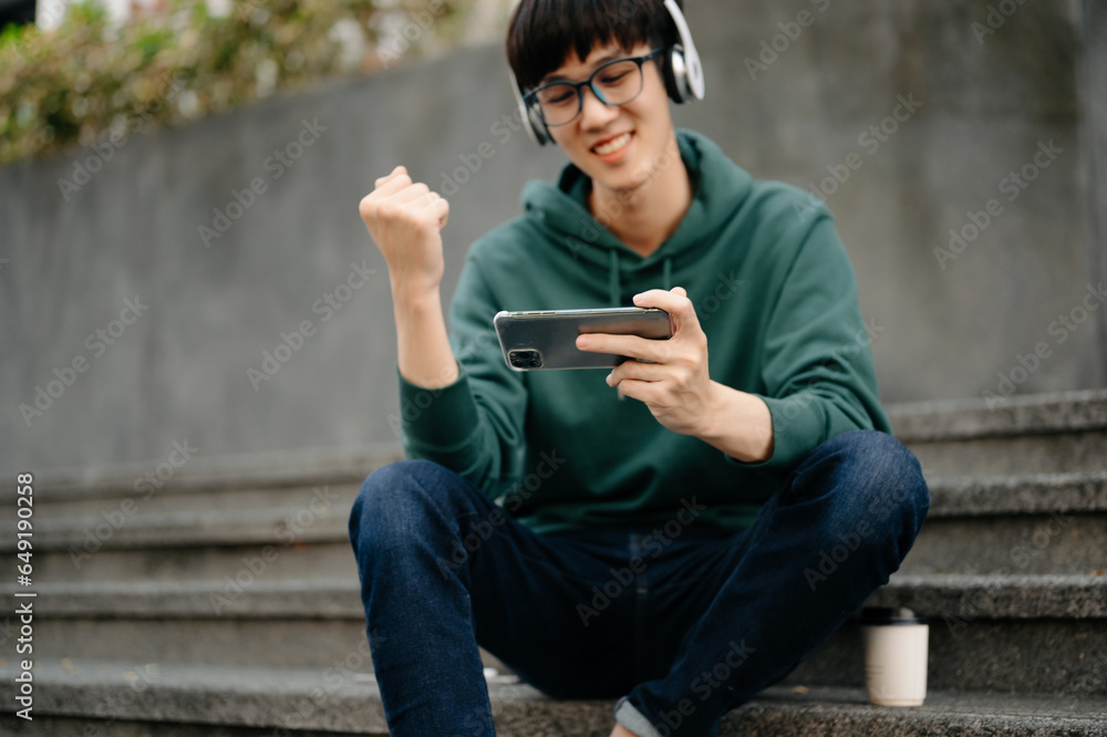 Young Asian man holds a cell phone in his hands typing messages or reading newsfeed.using modern gadget for entertainment playing different games online Young Asian man holds a cell phone in his hands typing messages or reading newsfeed.using modern gadget for entertainment playing different games online