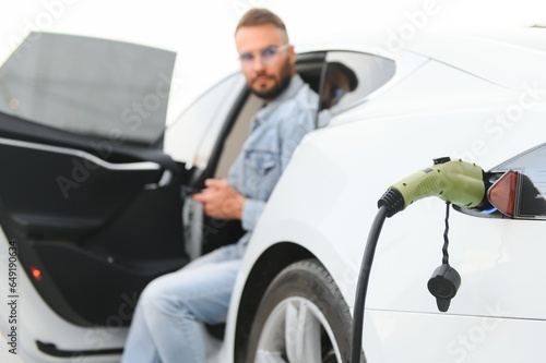Beautiful young stylish man is with electric car at daytime charging the vehicle